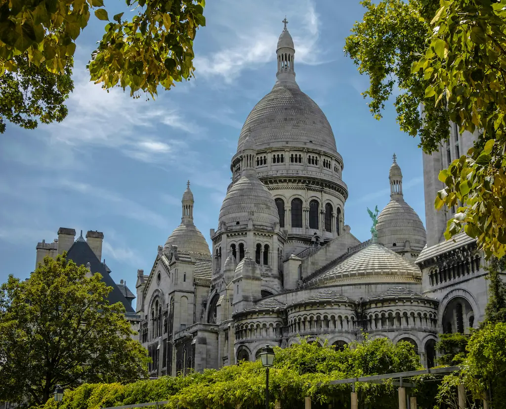 Kopuły i jasna elewacja bazyliki Sacré-Cœur na Montmartre, otoczone zielenią drzew.