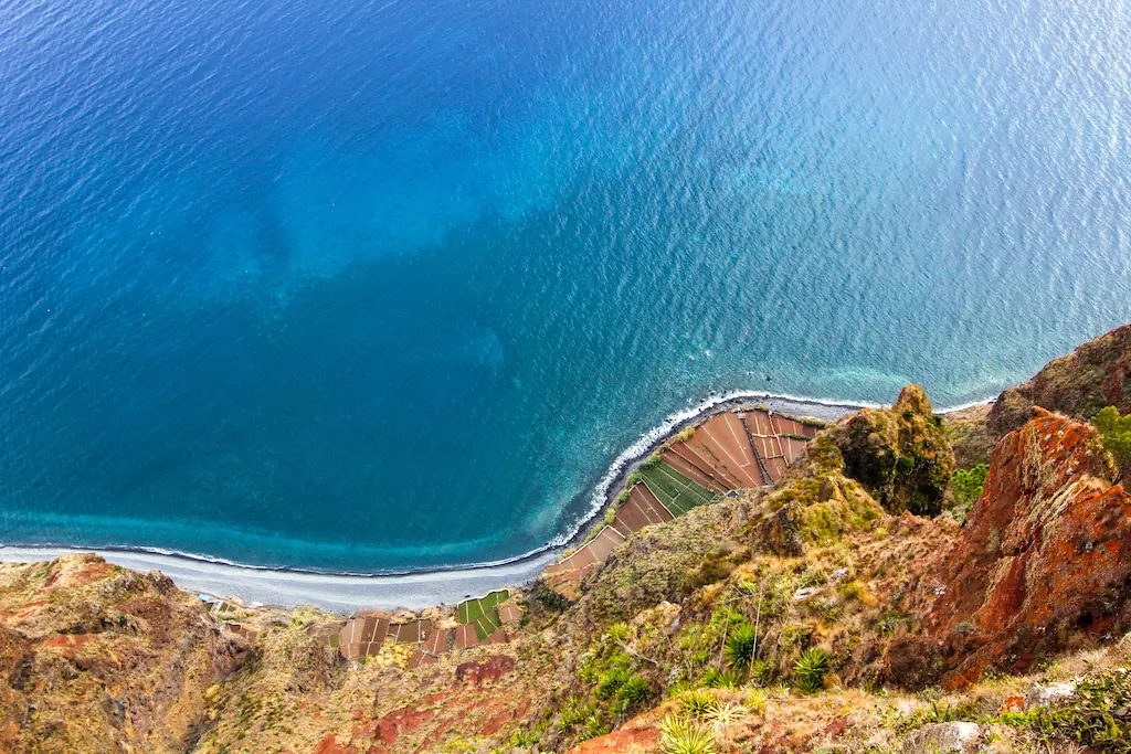 Widok z Cabo Girão na ocean, wąską plażę i pola u podnóża stromego klifu.