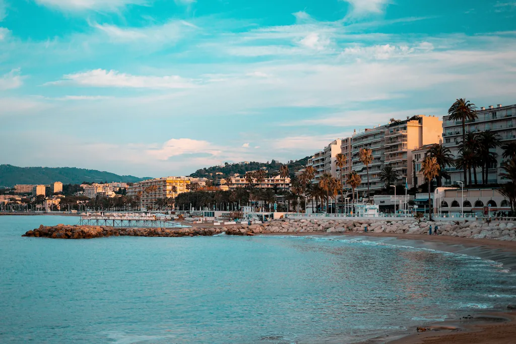 Plaża w Cannes z kamiennymi falochronami, palmami i zabudową przy Croisette.