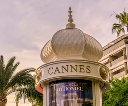 Ozdobna kopuła kiosku z napisem CANNES, otoczona palmami i nadmorską zabudową.