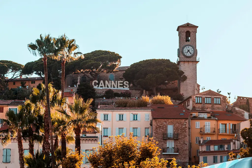 Wieża zegarowa nad kolorowymi domami w Cannes, z palmami i dużym napisem CANNES.