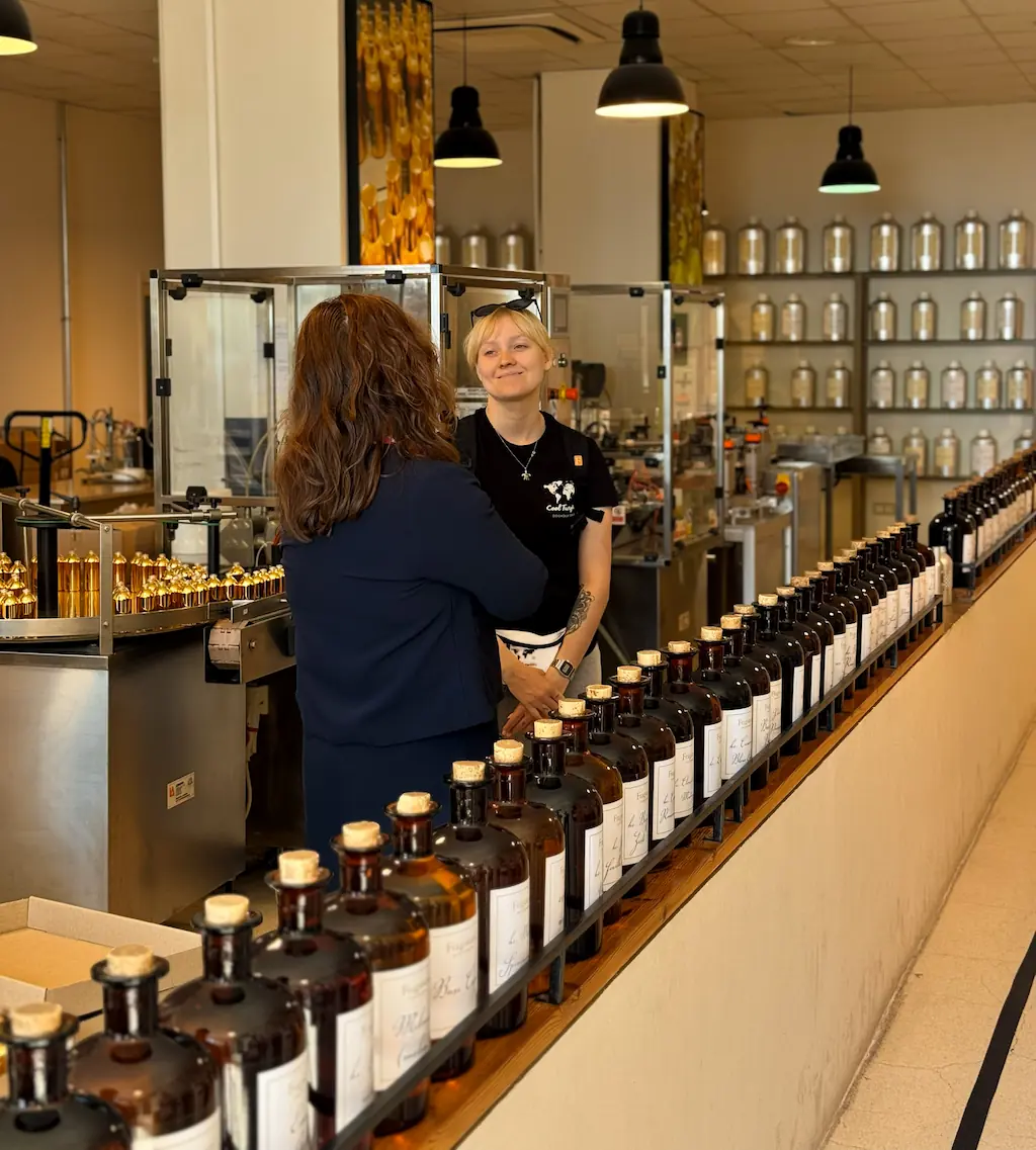 Uczestniczka wyjazdu CoolTurystek w fabryce perfum w Grasse, przy rzędzie brązowych butelek i urządzeniach produkcyjnych.