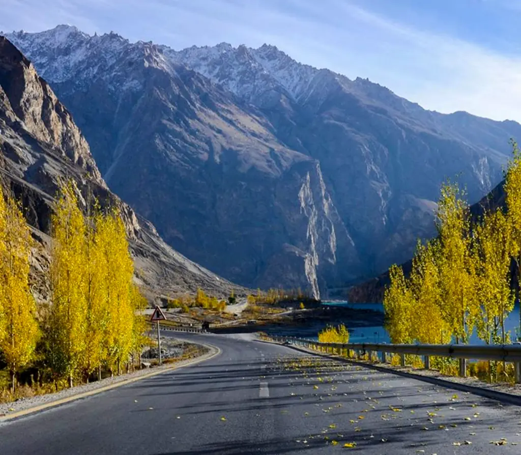 Zakręt Karakoram Highway między żółtymi drzewami, z barierą nad rzeką i stromymi skalistymi górami w tle
