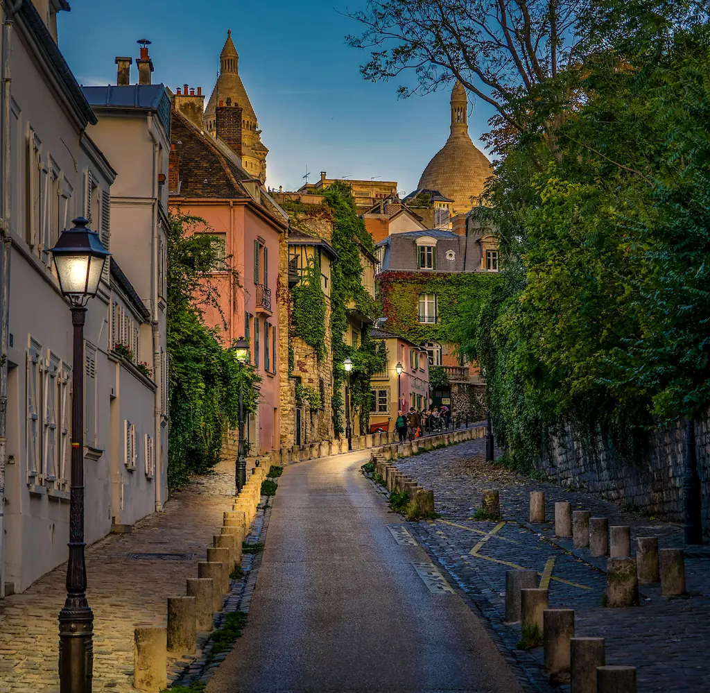 Brukowana uliczka na Montmartre z latarniami, kamienicami i kopułami Sacré-Cœur ponad dachami.