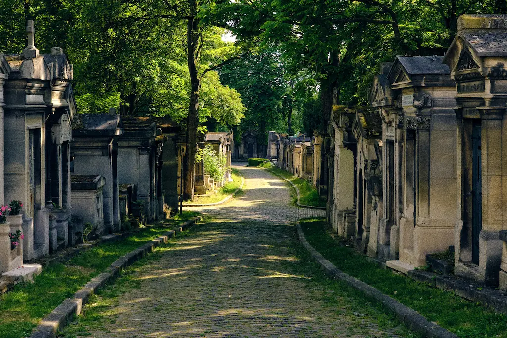 Brukowana aleja cmentarza Père-Lachaise pomiędzy kamiennymi grobowcami i wysokimi drzewami.