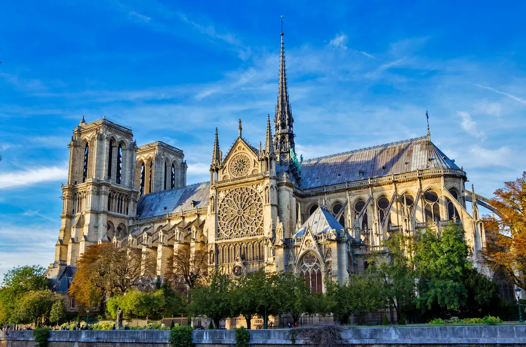 Katedra Notre-Dame widziana znad Sekwany, z wieżami, rozetą i iglicą ponad otaczającymi ją drzewami.
