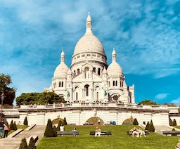 Fasada bazyliki Sacré-Cœur ponad schodami i zielonym zboczem wzgórza Montmartre.